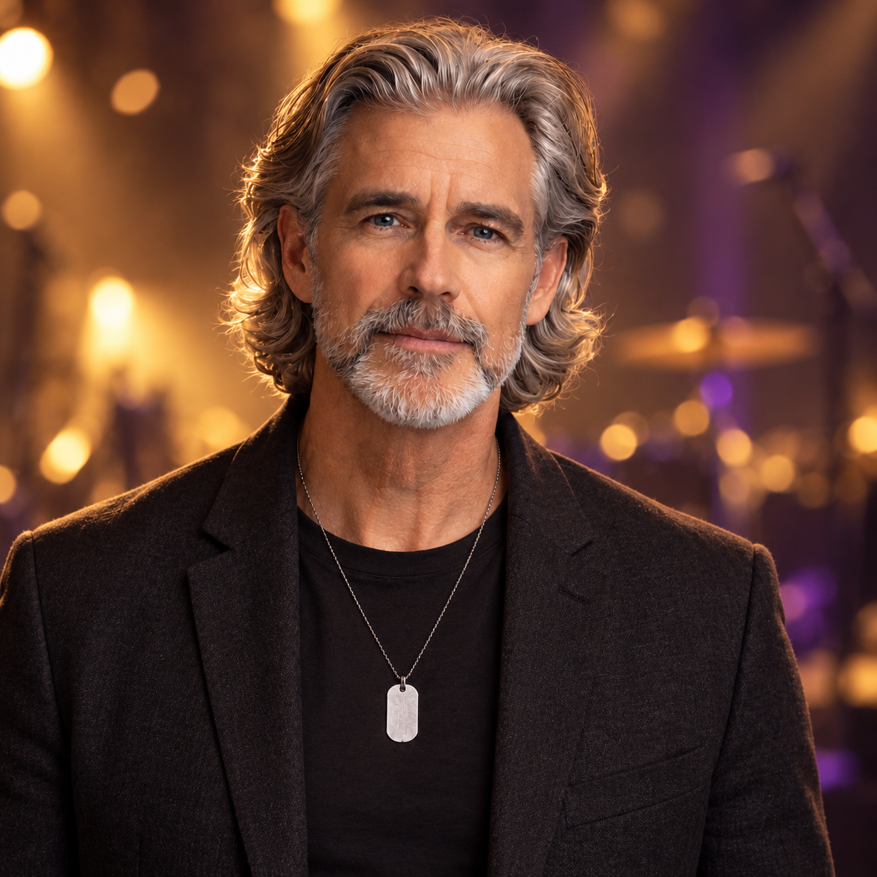 David in a black shirt and charcoal jacket with his silver dog-tag necklace, against softly blurred amber and purple stage lights